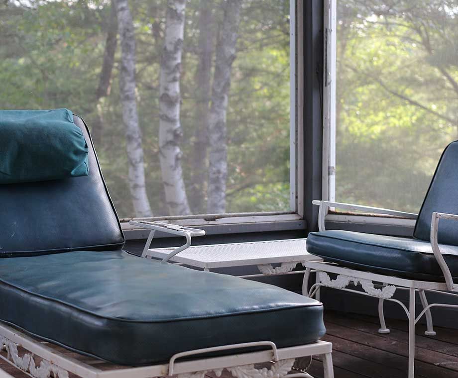 Une véranda avec une méridienne et un fauteuil bleus, une table blanche, donnant sur des bouleaux.