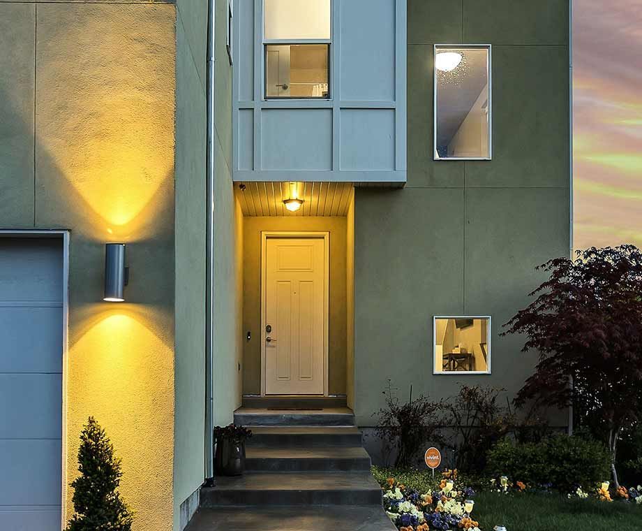 Maison moderne à deux étages avec façade verte. Porte d'entrée visible avec escalier menant à l'étage, éclairée par une lumière chaude.