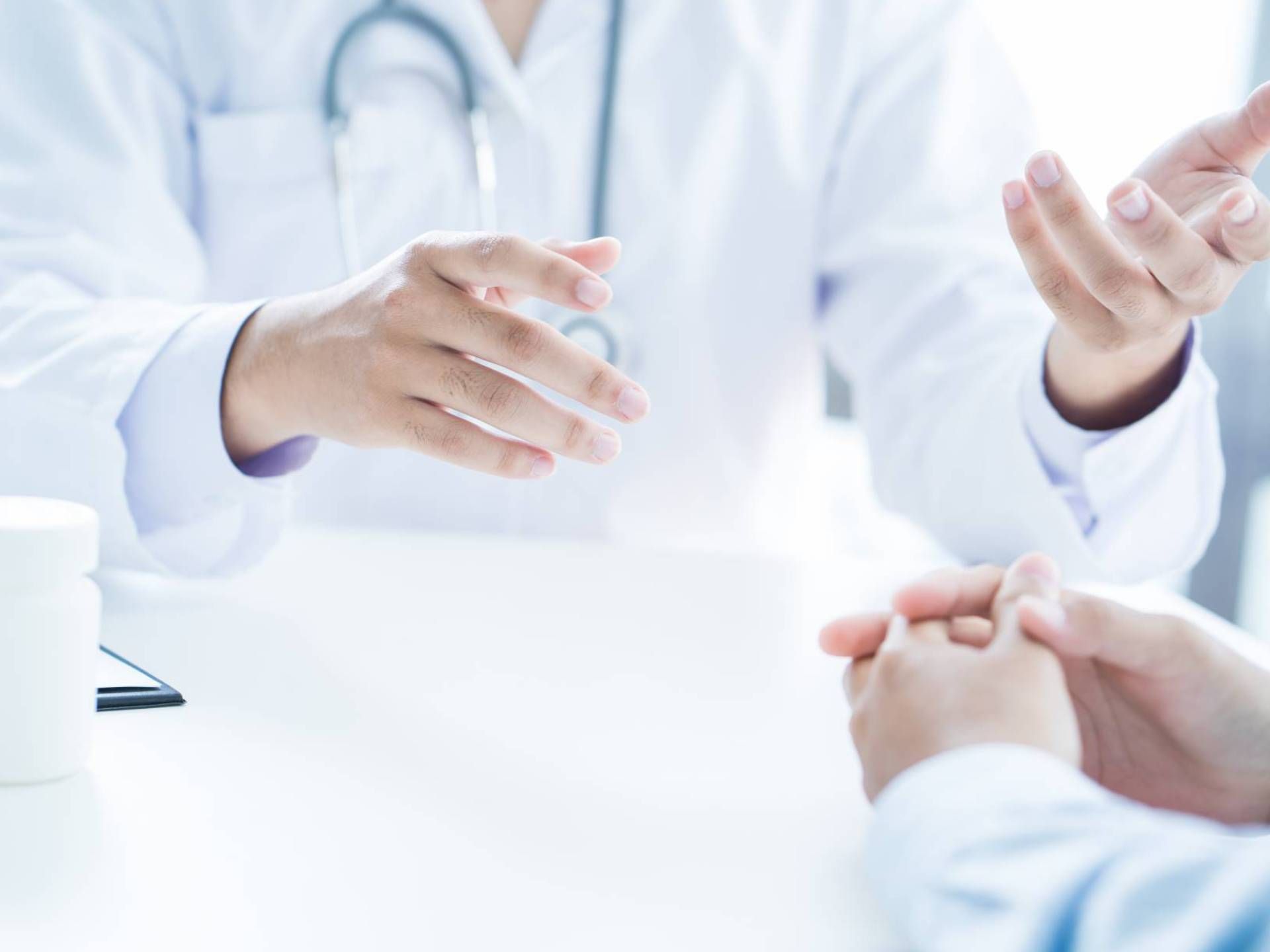 Person in weißem Kittel mit Stethoskop gestikuliert vor einer Person mit gefalteten Händen