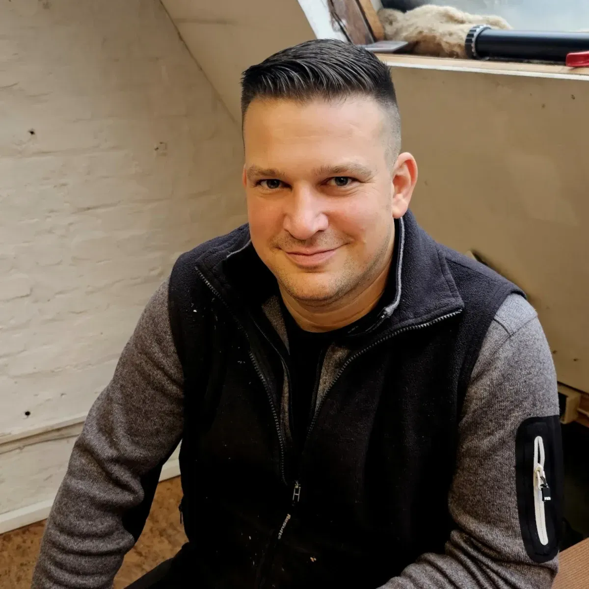 Eine Person sitzt drinnen, trägt eine schwarze Weste und einen grauen Pullover und lächelt leicht.