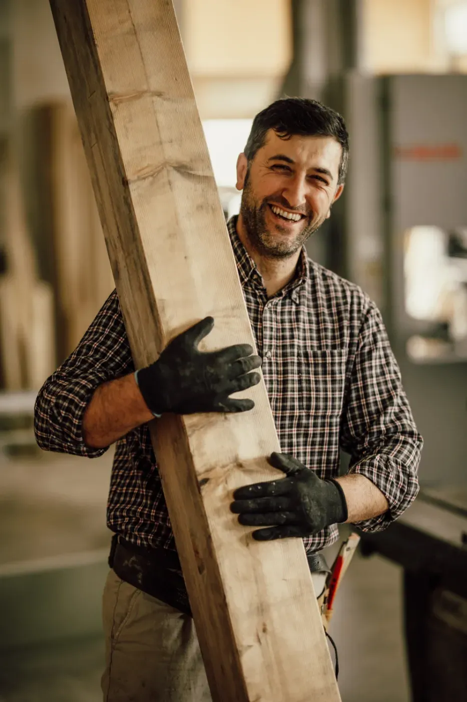 Ein lächelnder Arbeiter mit Handschuhen trägt einen großen Holzbalken in einer Werkstatt.