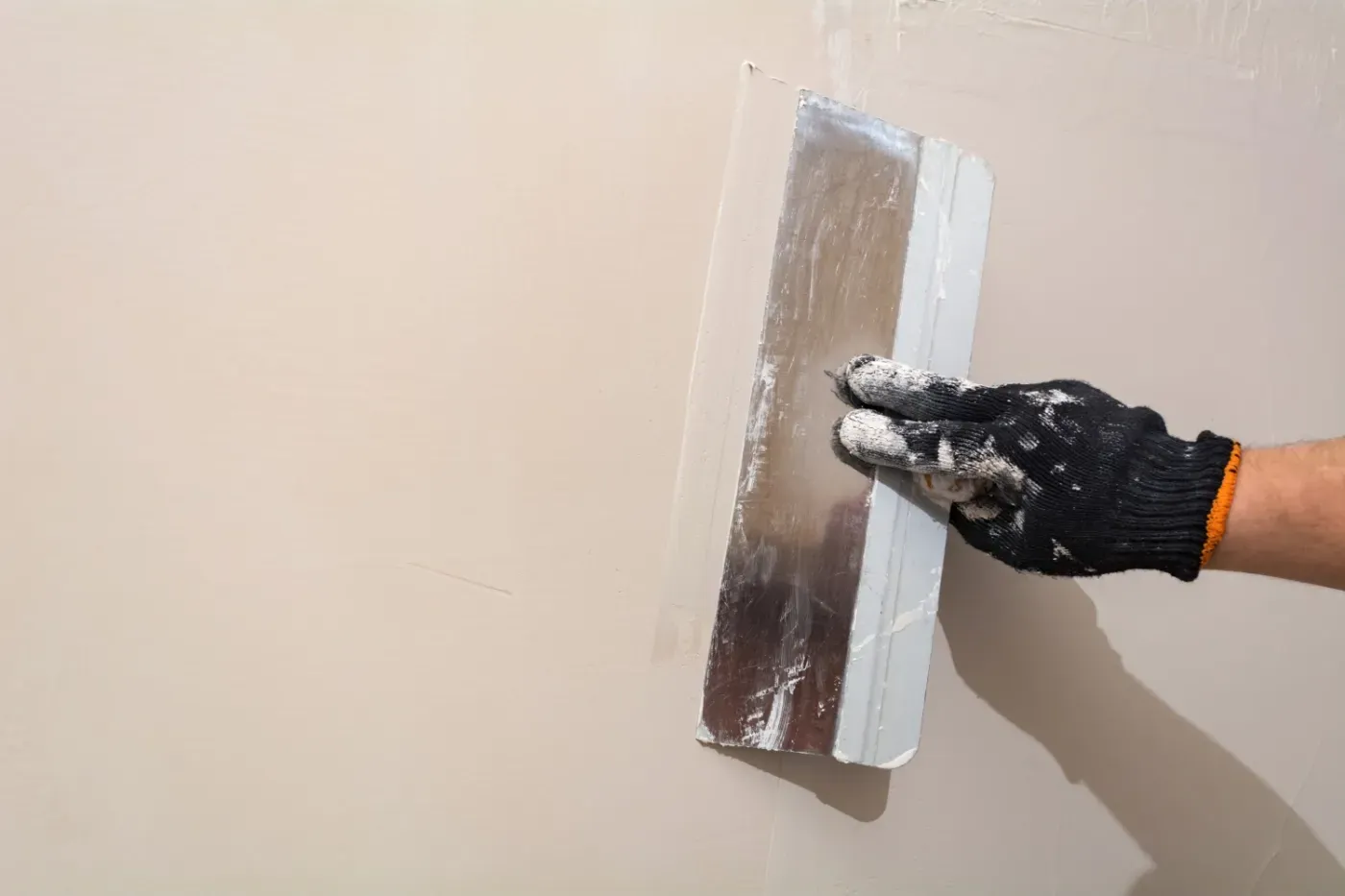 Eine behandschuhte Hand glättet mit einer Kelle Putz auf einer beigen Wand