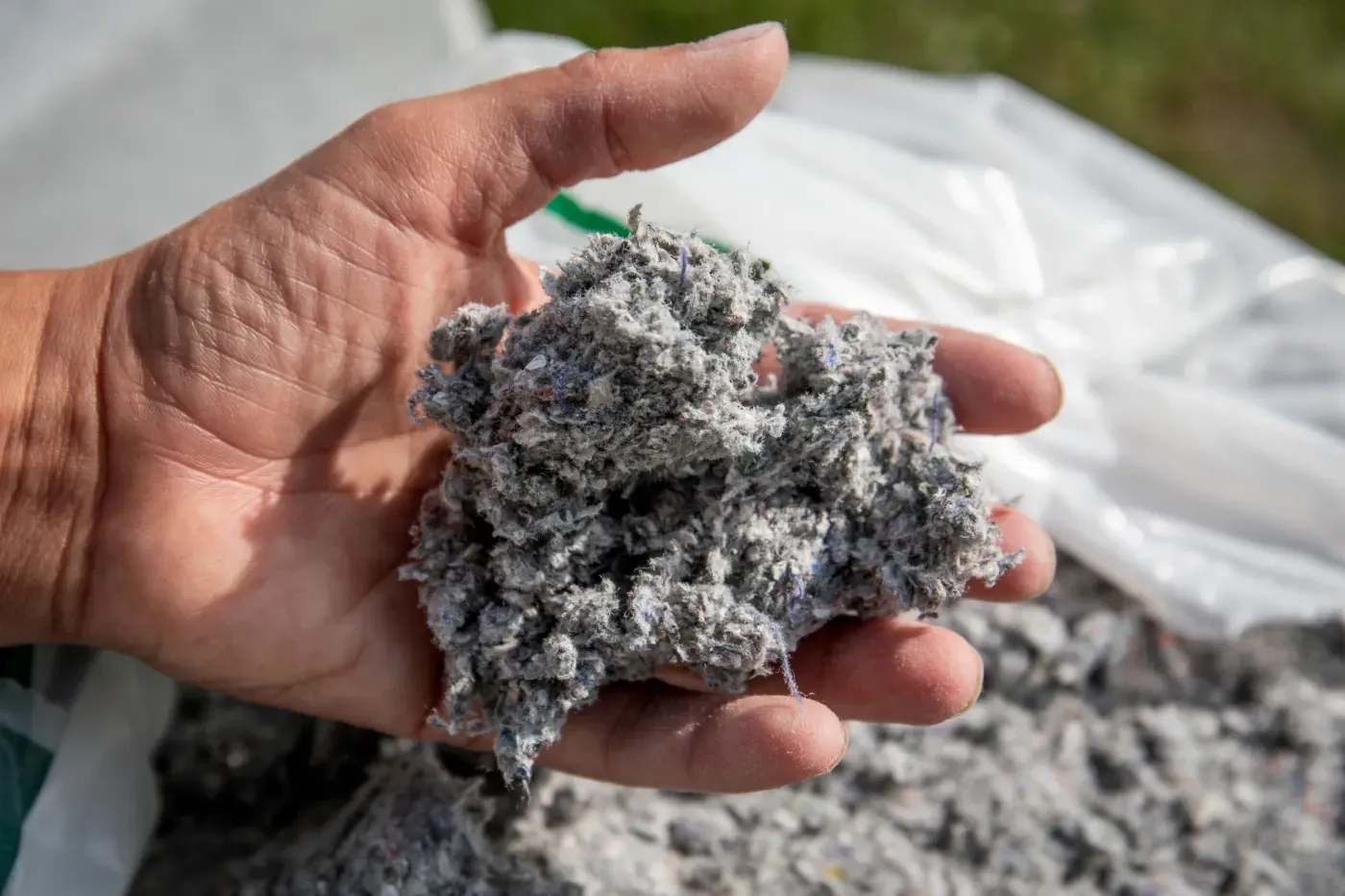 Eine Hand hält im Freien einen grauen Klumpen faserigen Materials, im Hintergrund ist weiteres Material aufgestapelt.