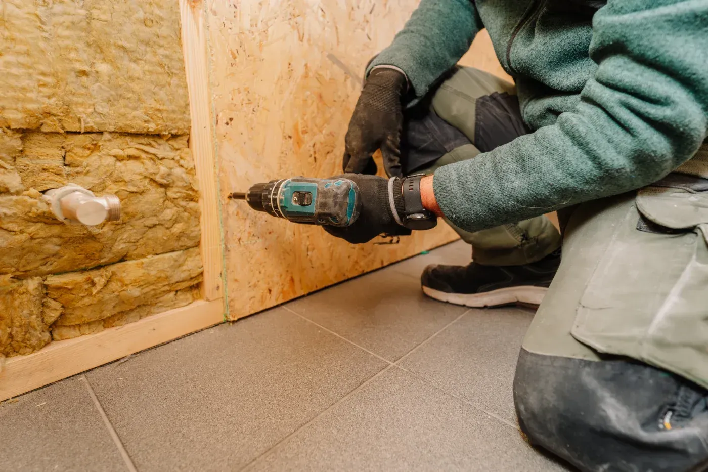 Eine Person benutzt eine Bohrmaschine, um Holzpaneele an einer Wand auf einem gefliesten Boden anzubringen.