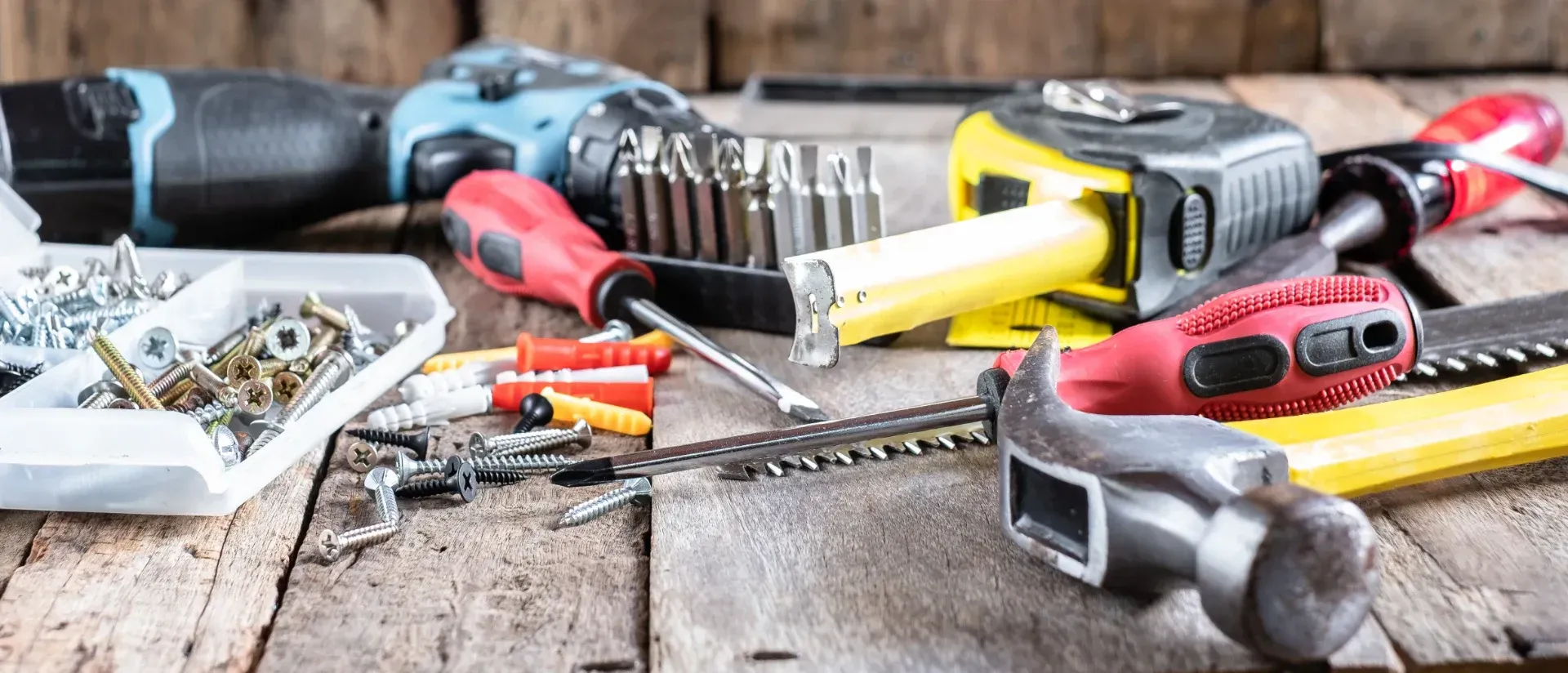 Verschiedene Handwerkzeuge, Bohrmaschine, Hammer, Schraubenschlüssel, Schraubendreher und Schrauben liegen auf einer hölzernen Werkbank verstreut.