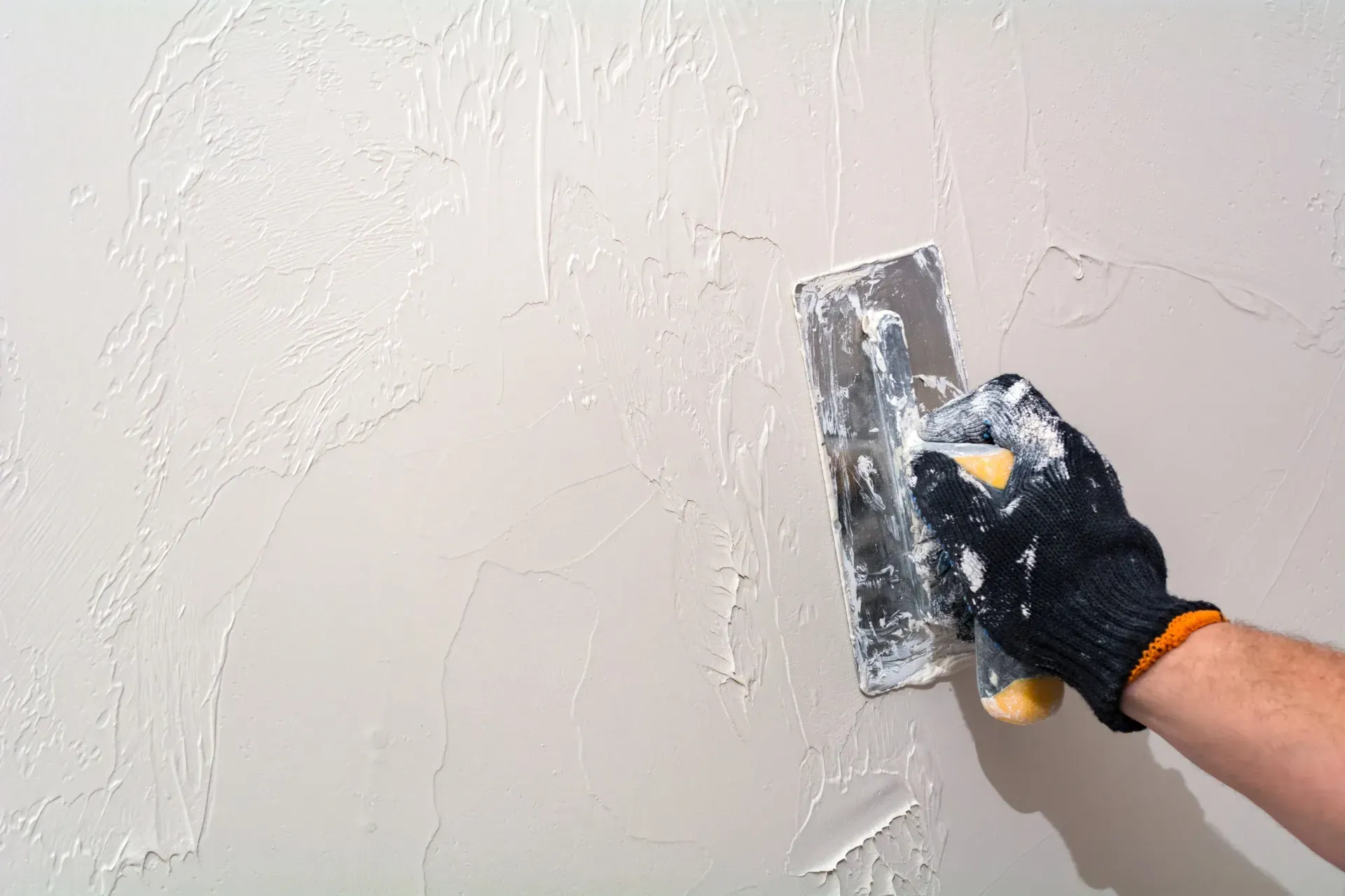 Eine behandschuhte Hand glättet mit einer Kelle weißen Putz an einer Wand.