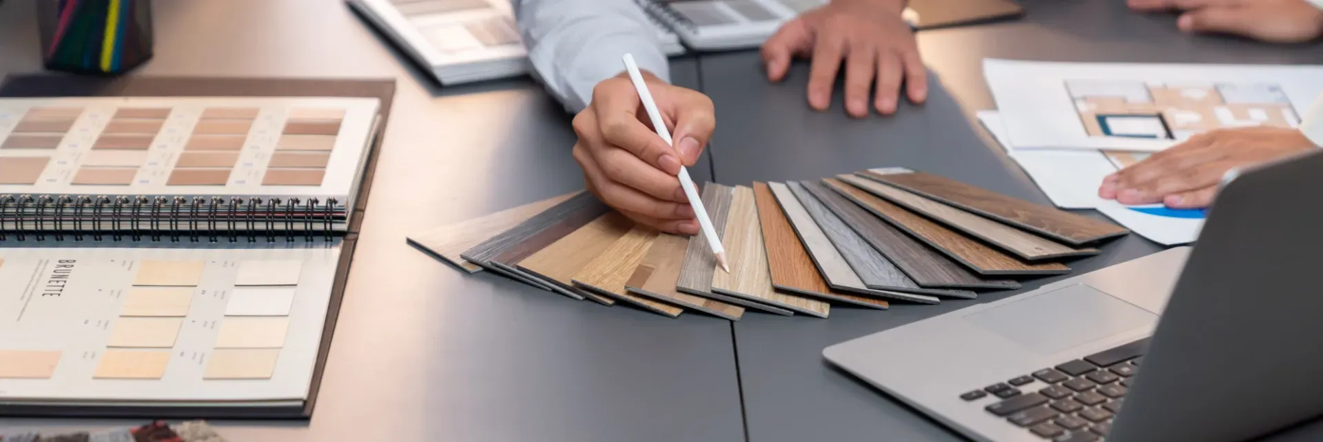 Hände betrachten Stoffmuster und Farbproben auf einem Designtisch, Laptop und Dokumente liegen daneben.