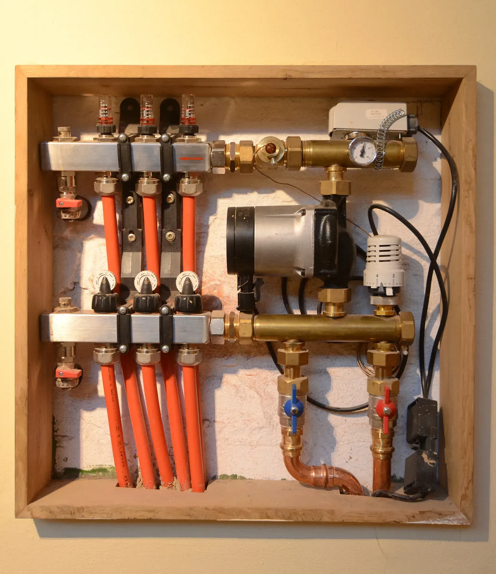 Offener Wandkasten mit Verteilerkasten, Ventilen, Rohren und einer kleinen Pumpeneinheit in einem Hauswirtschaftsraum.