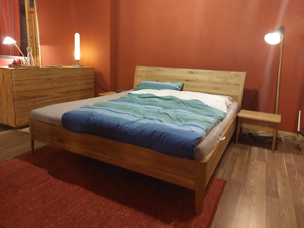 Ein Holzbett in einem Zimmer mit roter Wand und Teppich. Es hat eine blau-grüne Decke und steht neben einem kleinen Nachttisch.