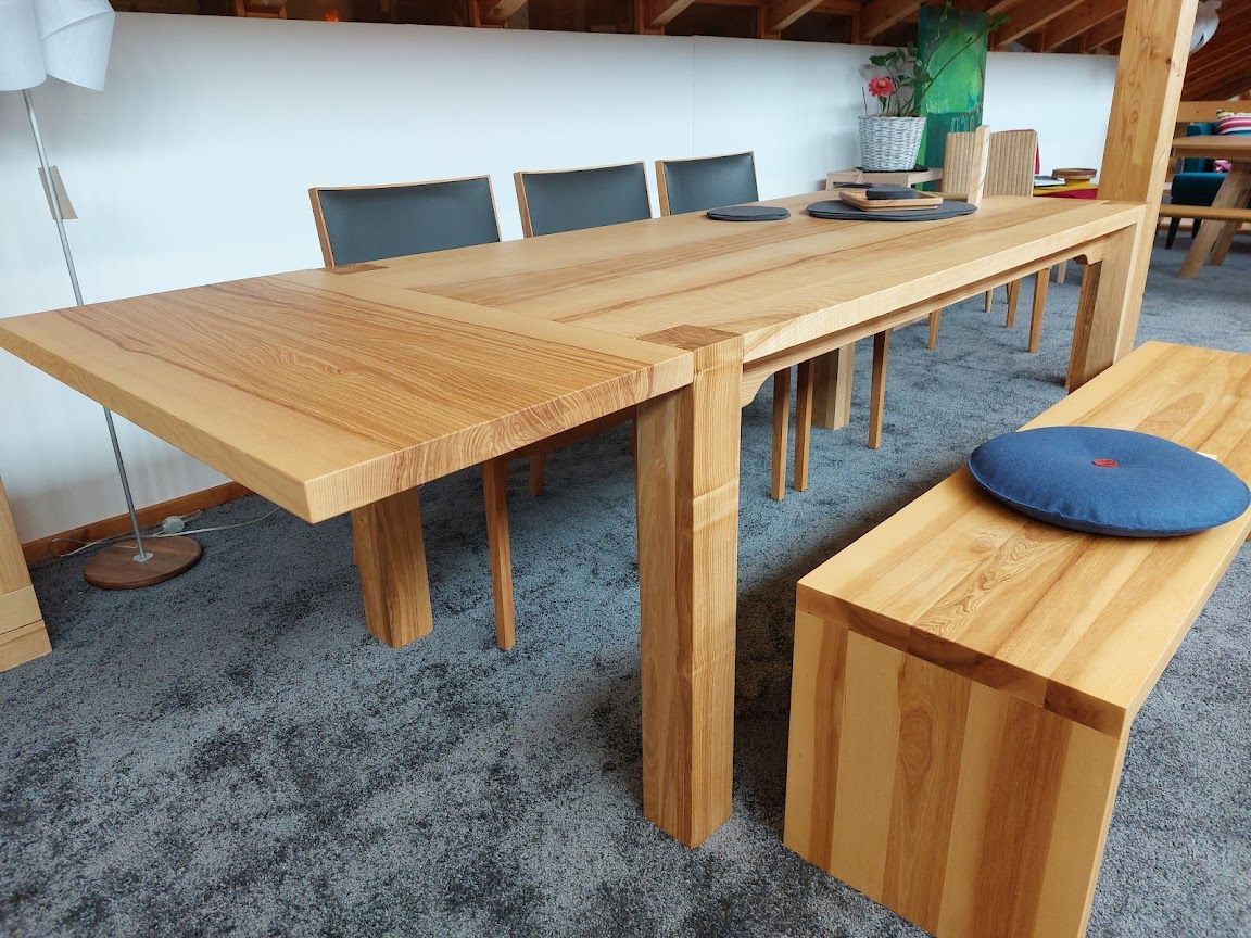 Ein Esstisch aus Holz mit Bank und Stühlen steht vor einer weißen Wand in einem Zimmer mit grauem Teppichboden.