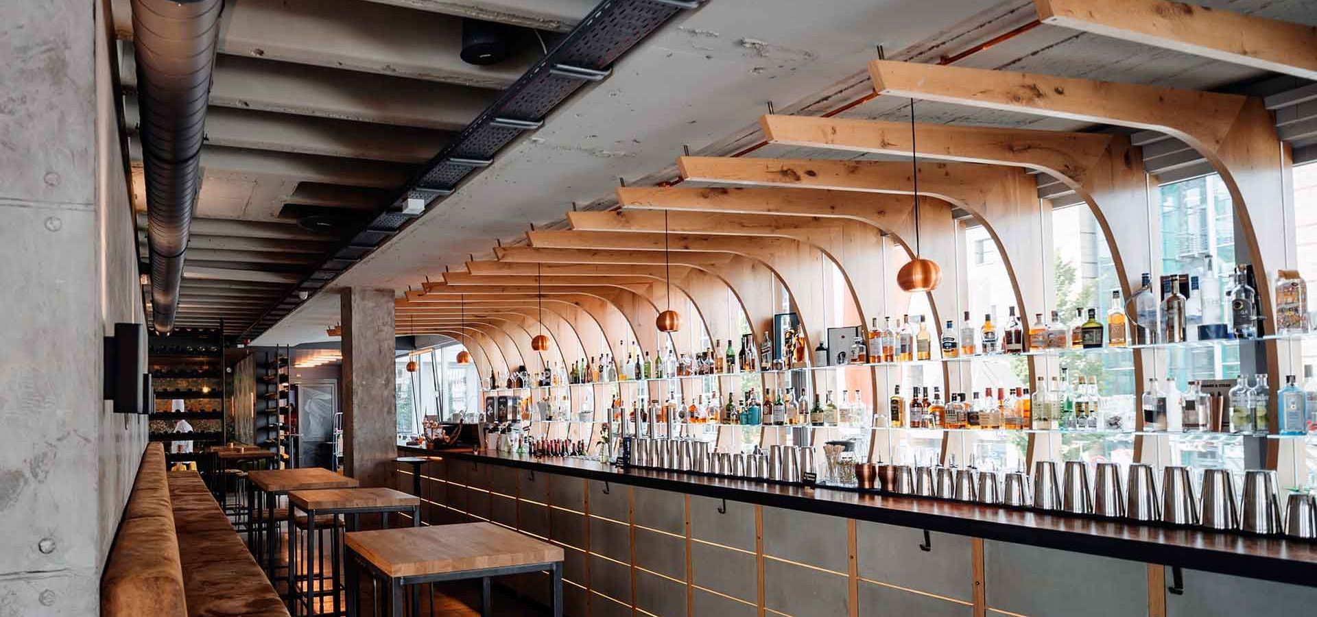Innenansicht einer Bar mit hölzerner Deckengestaltung und einer langen Theke, an der Flaschen und Shaker ausgestellt sind.