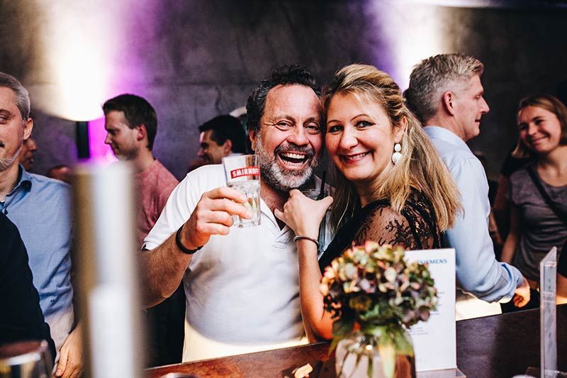 Menschen bei einer Zusammenkunft, lächelnd, einer hält ein Getränk in der Hand.