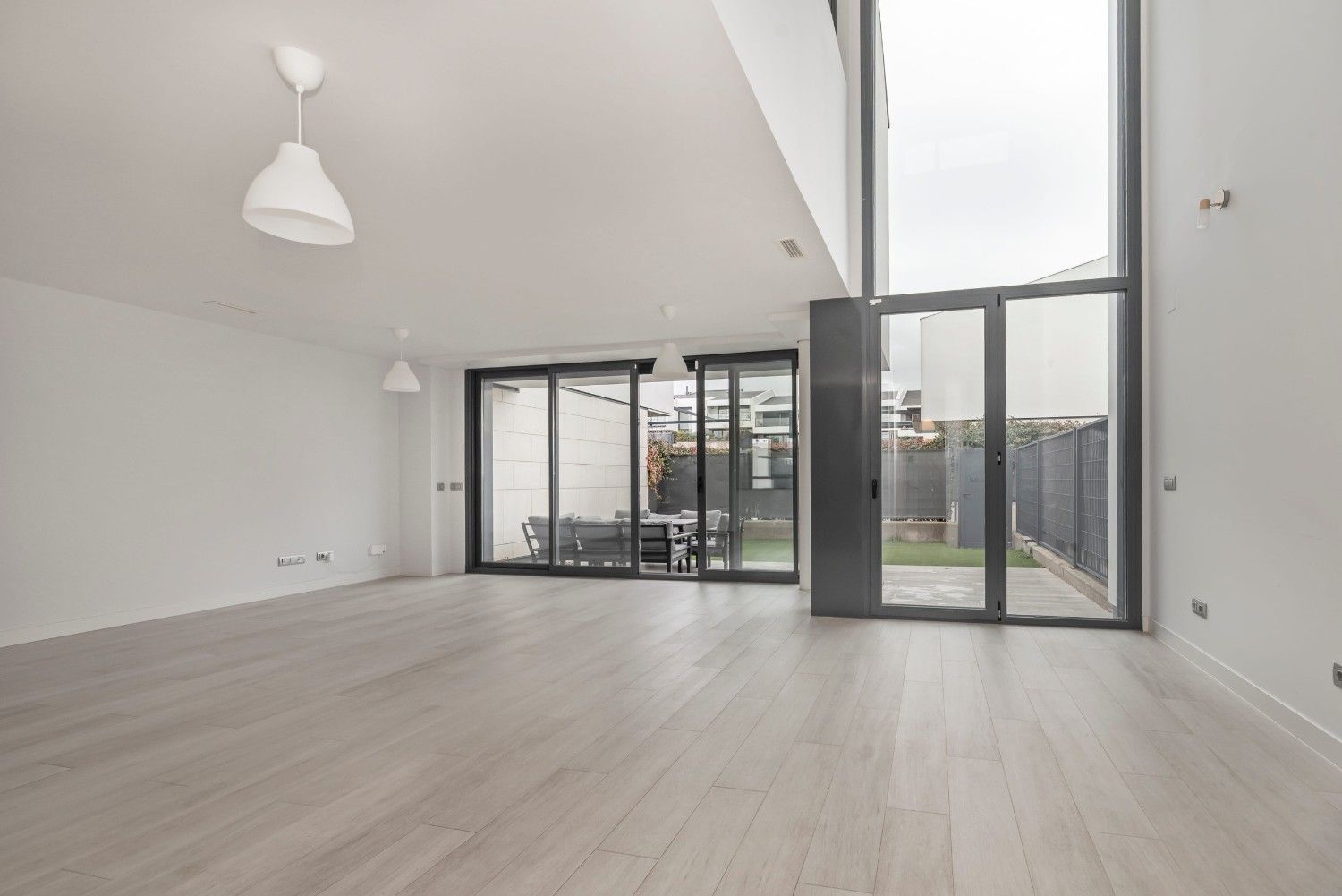 Amplio salón moderno y vacío con suelos de madera clara, paredes blancas y grandes puertas de cristal que dan a un patio.