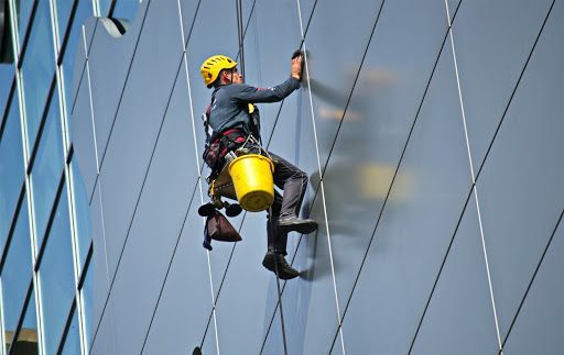 Limpiador de ventanas con casco amarillo y equipo de seguridad desciende en rápel por la fachada de cristal de un edificio.