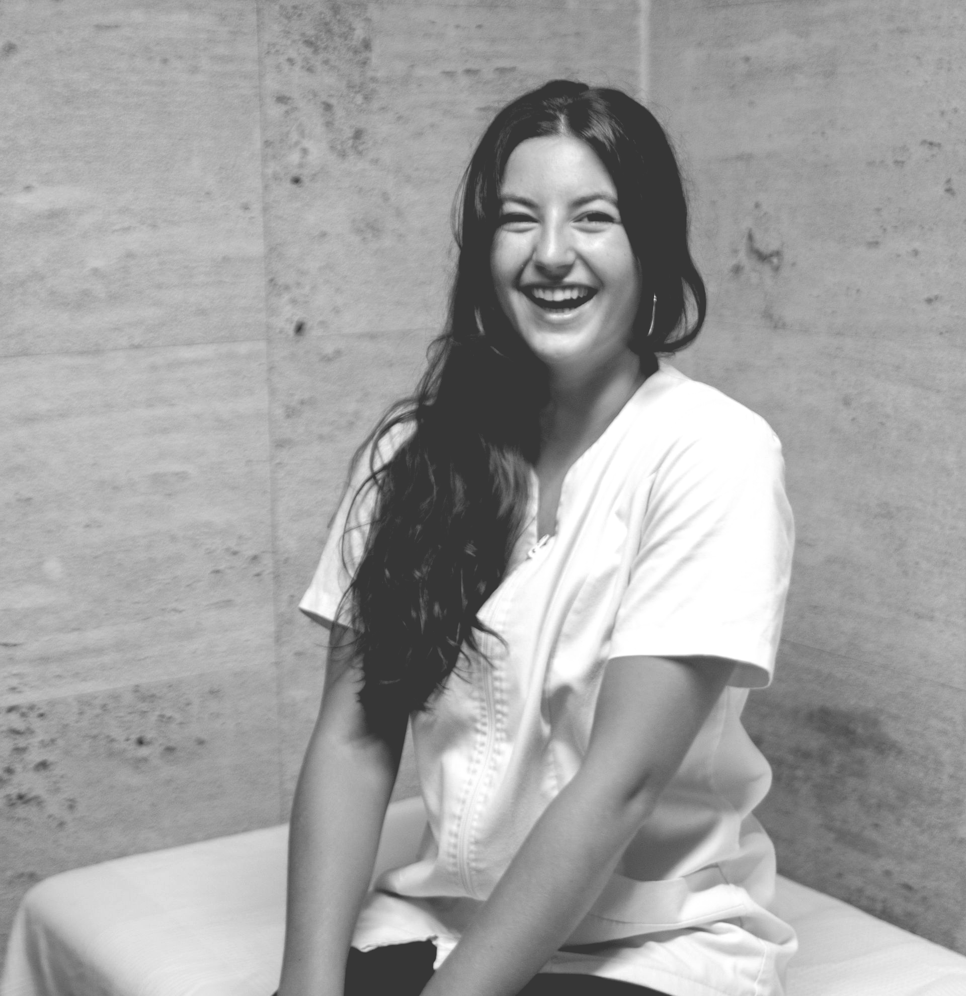 Una mujer vestida con uniforme blanco ríe, sentada en una camilla de masaje, apoyada contra una pared de mármol.