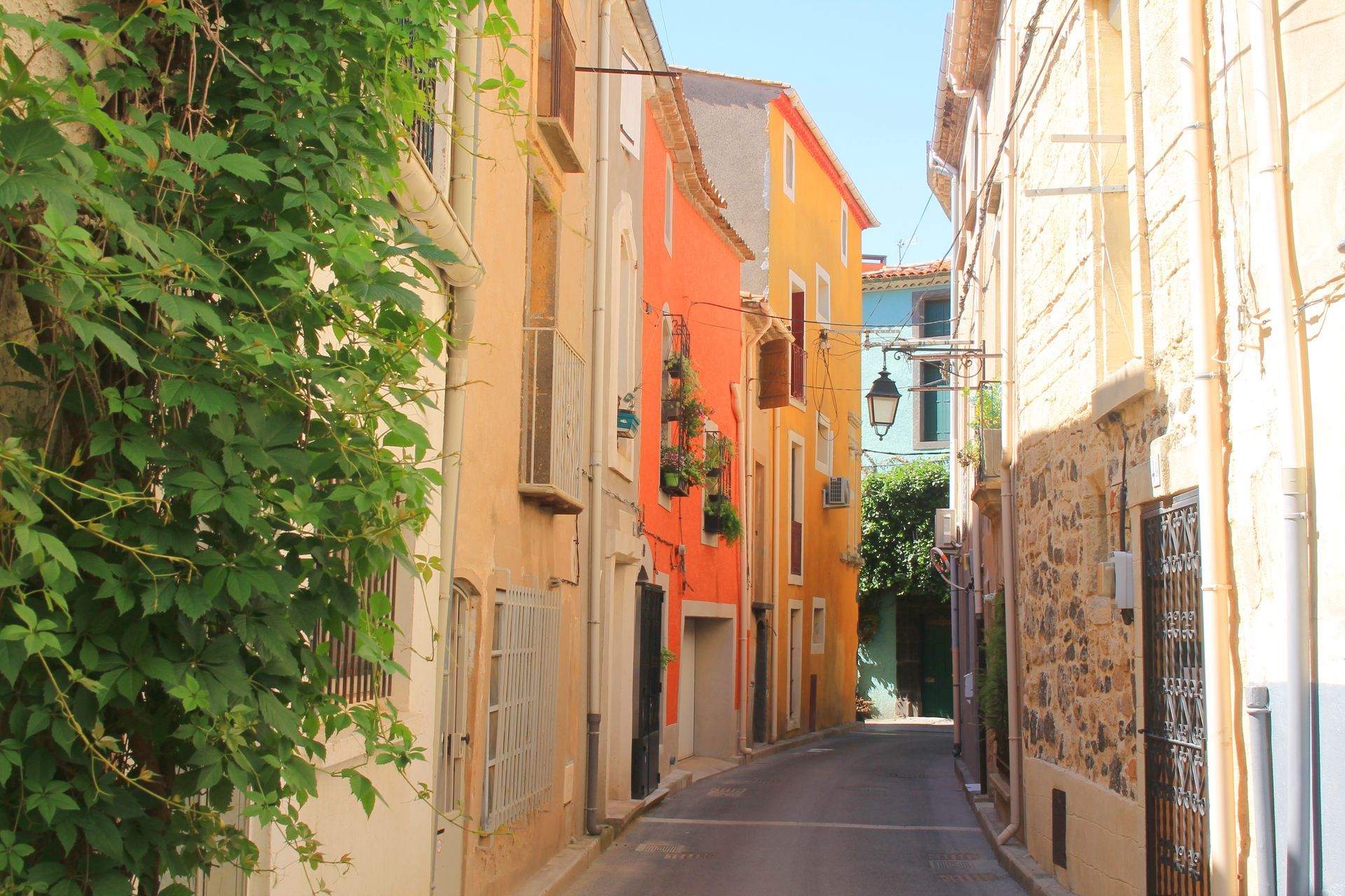 Ruelle étroite et ensoleillée bordée de bâtiments colorés, de lierre grimpant et d'une arche bleue que l'on aperçoit au loin.