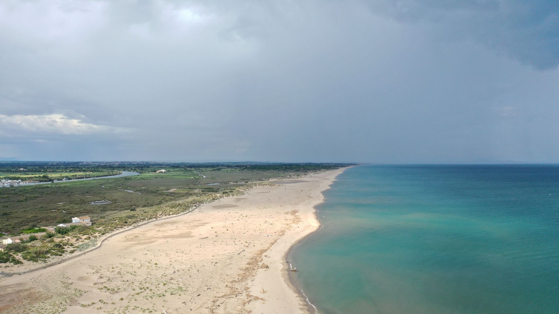 Vue aérienne d'une plage de sable fin qui s'incurve au bord d'un océan turquoise sous un ciel nuageux.