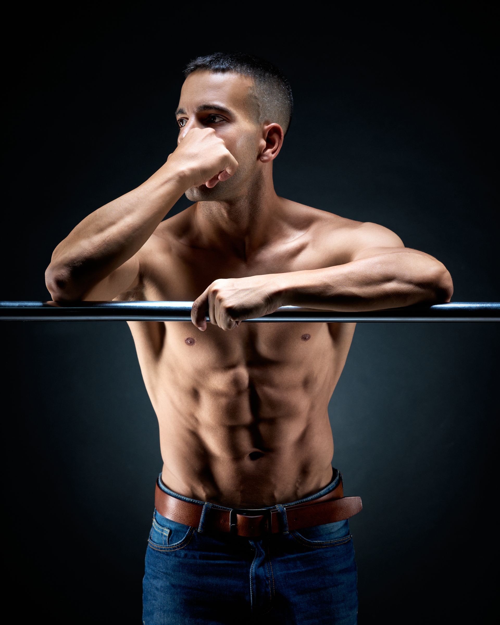 Hombre en forma, sin camisa, apoyado en una barra, con vaqueros. Parece pensativo, con la mano cerca de la cara, fondo oscuro