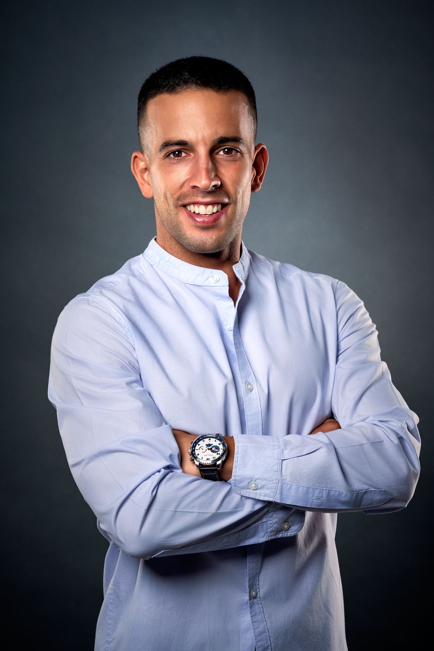 Hombre con los brazos cruzados, con camisa azul claro, sonriendo a la cámara. Fondo oscuro.