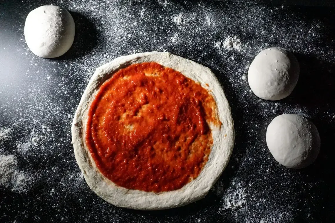 Una pizza con salsa de tomate encima está sobre una mesa rodeada de bolas de masa.