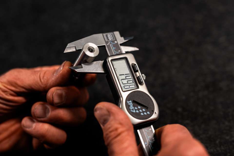 Person misst den Durchmesser einer Schraube mit einem digitalen Messschieber, schwarze Einstellung, Messwert 14,98 mm.