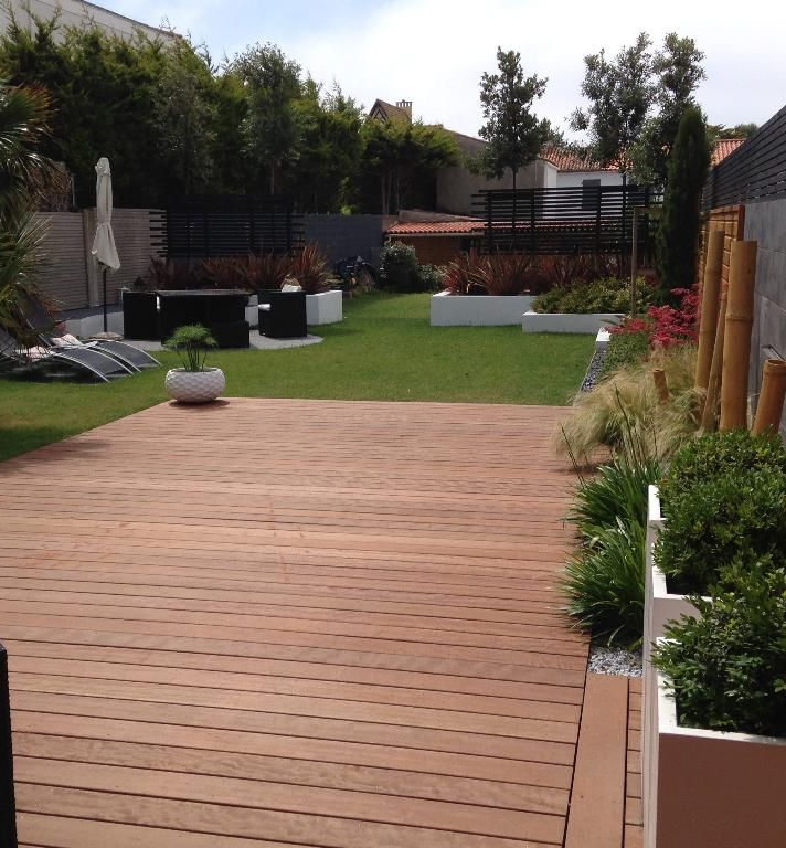 Une terrasse en bois donne sur un jardin luxuriant avec pelouse, plantes et espaces de détente.