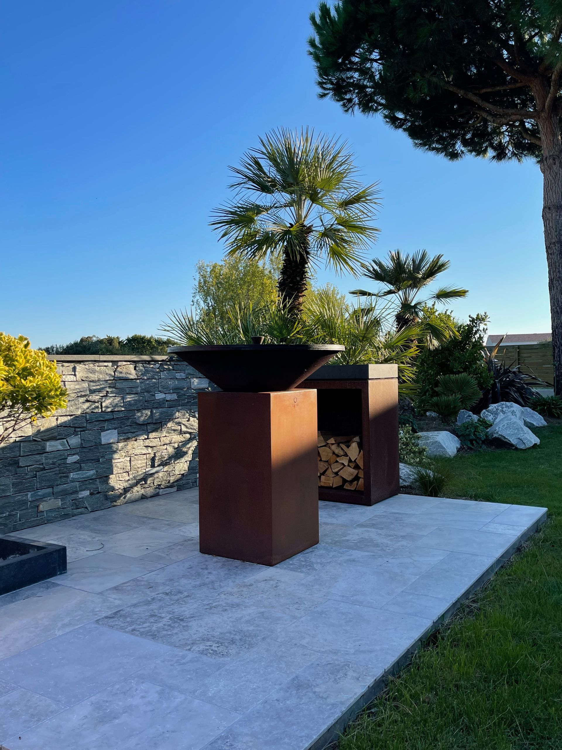 Foyer extérieur avec rangement pour le bois de chauffage sur une terrasse en béton, près d'un mur de pierre et de verdure, en plein soleil.