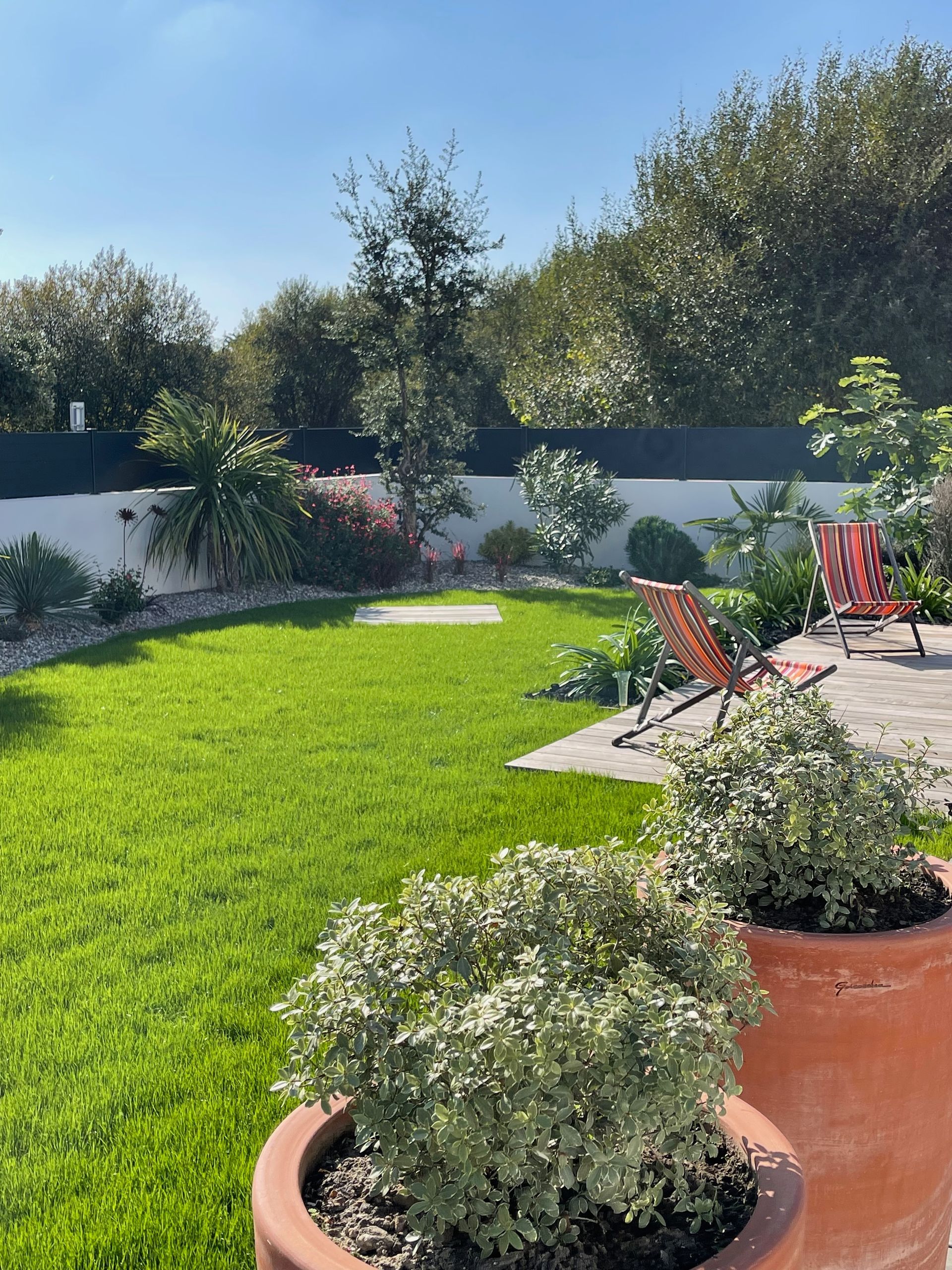 Pelouse verte avec parterres de fleurs, arbres et deux chaises orange dans un jardin ensoleillé.