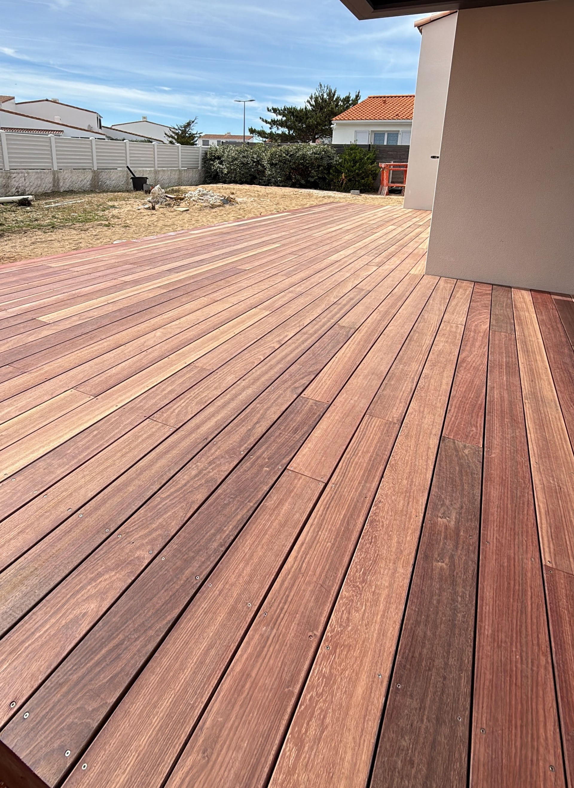 Terrasse en bois ensoleillée, avec le mur de la maison à droite et un jardin clairsemé en arrière-plan.