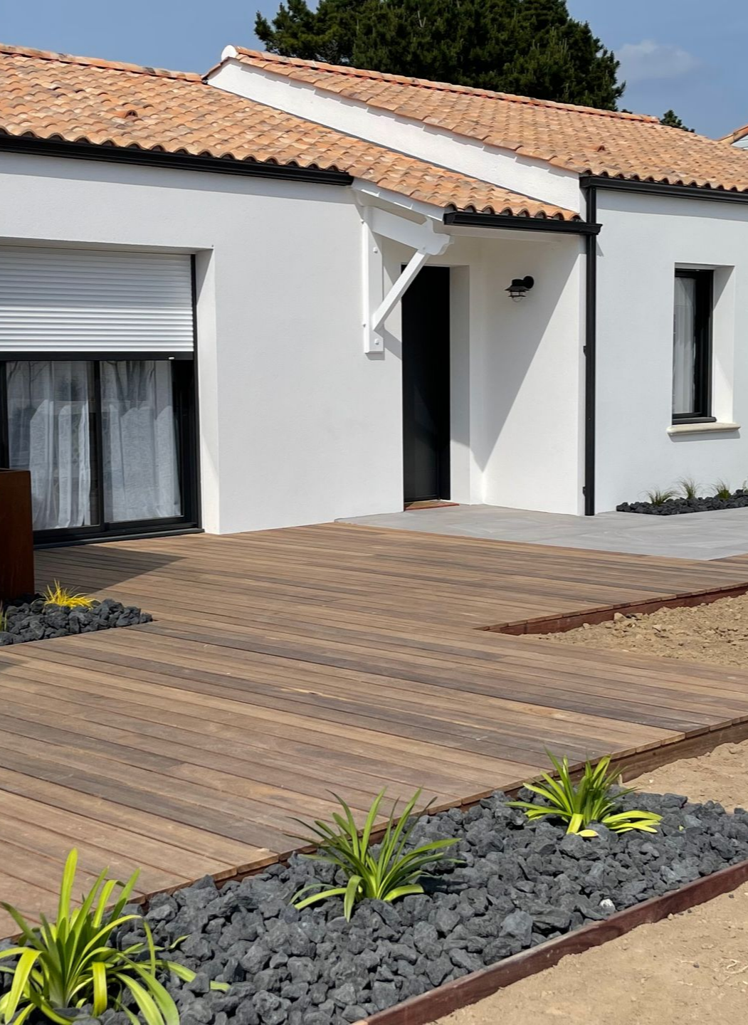 Maison blanche avec toit en tuiles de terre cuite, encadrements de fenêtres et porte noirs, terrasse en bois et aménagement paysager.