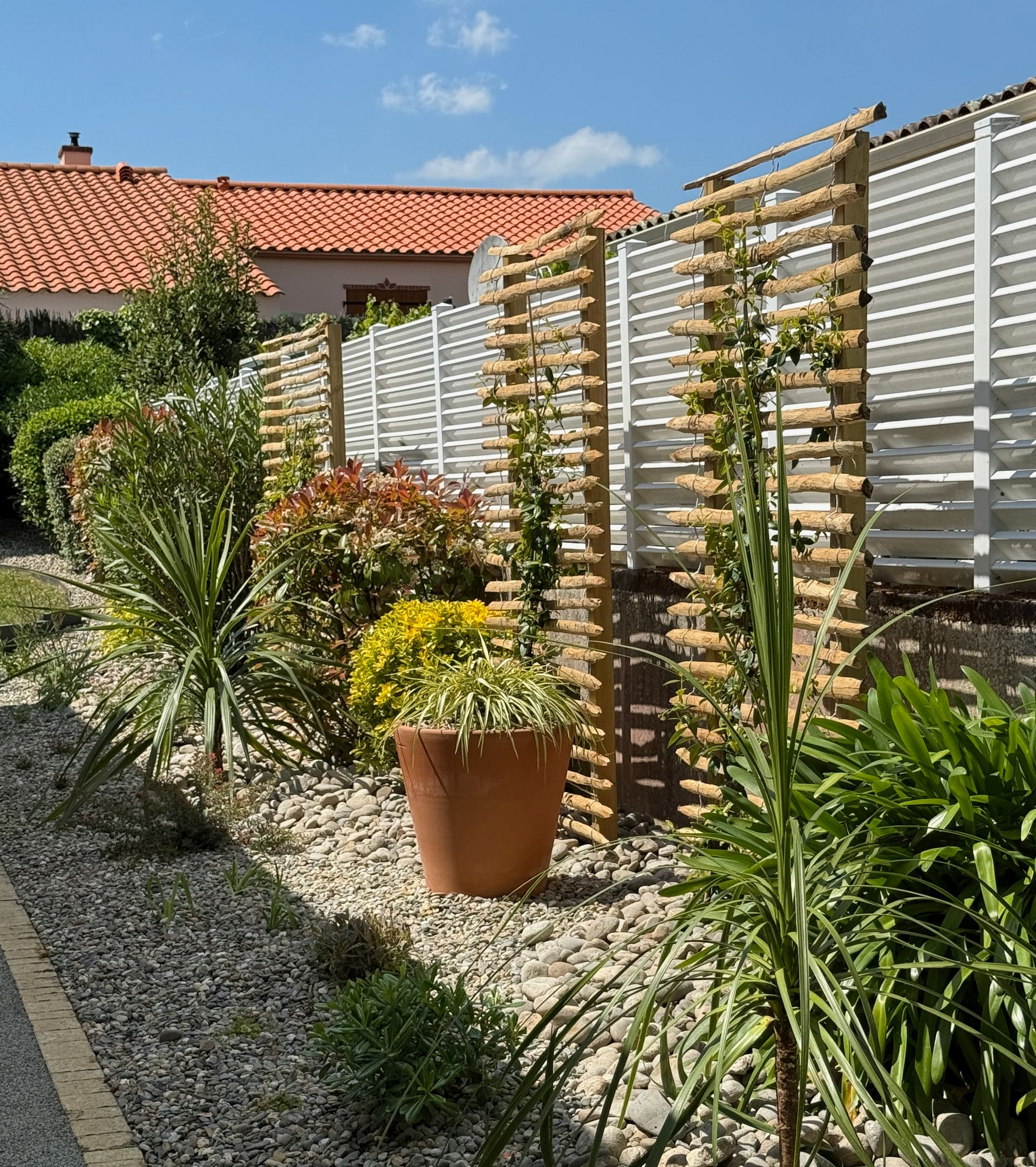 Un parterre de jardin avec des plantes et un treillis en bois se trouve devant une clôture blanche ; un pot en terre cuite est visible.