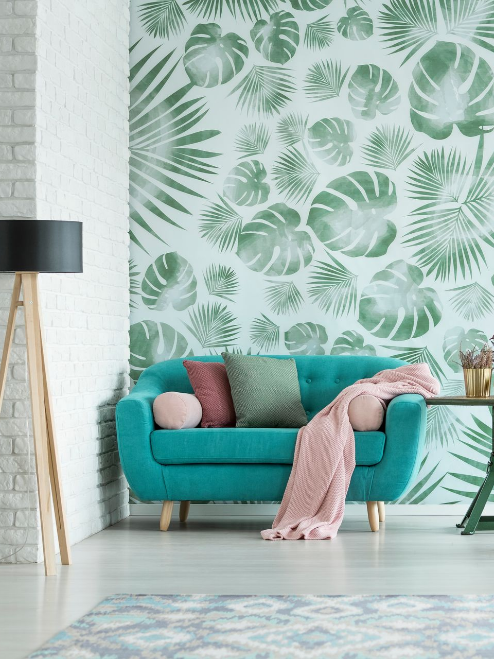 Canapé bleu sarcelle avec jeté et coussins roses devant un papier peint à feuilles tropicales, près d'une lampe en bois.