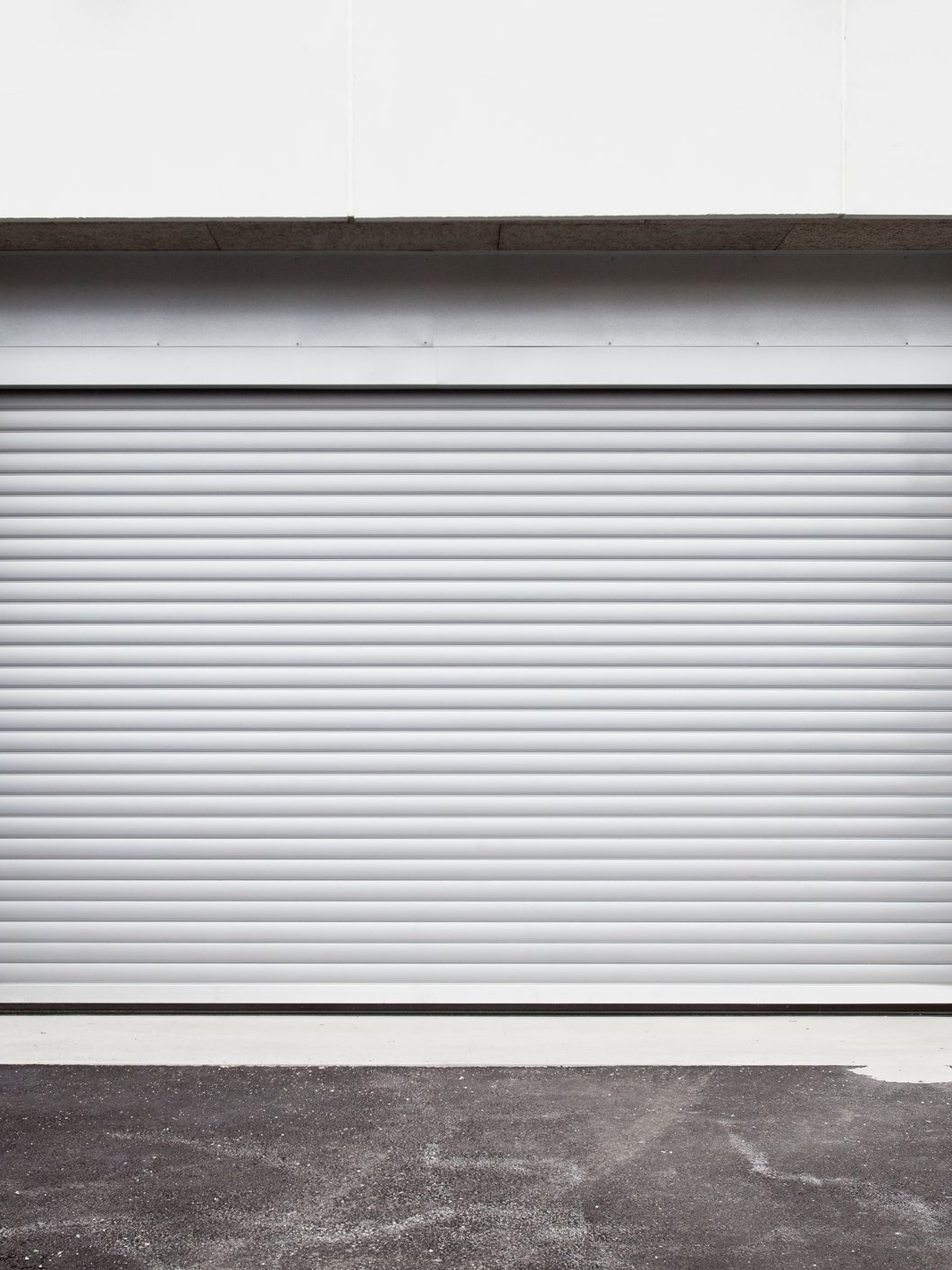 Porte de garage enroulable grise et fermée devant un bâtiment blanc, sur une surface en béton.