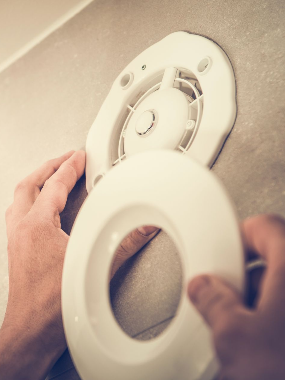 Mains installant un couvercle de ventilateur d'extraction de salle de bain blanc sur un mur de couleur claire.