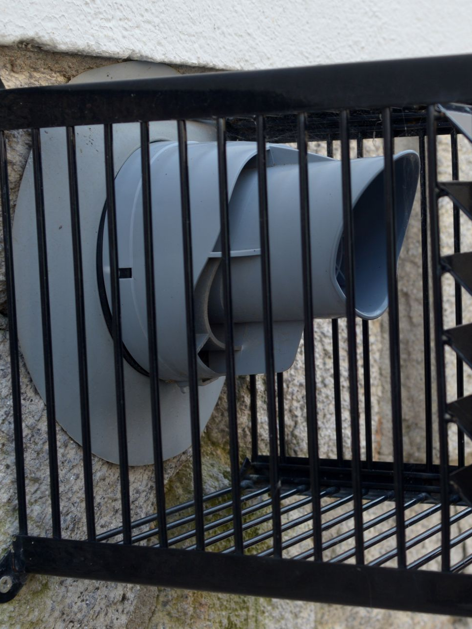 Évent d'échappement gris avec une cage de protection en métal noir sur un extérieur de bâtiment blanc.