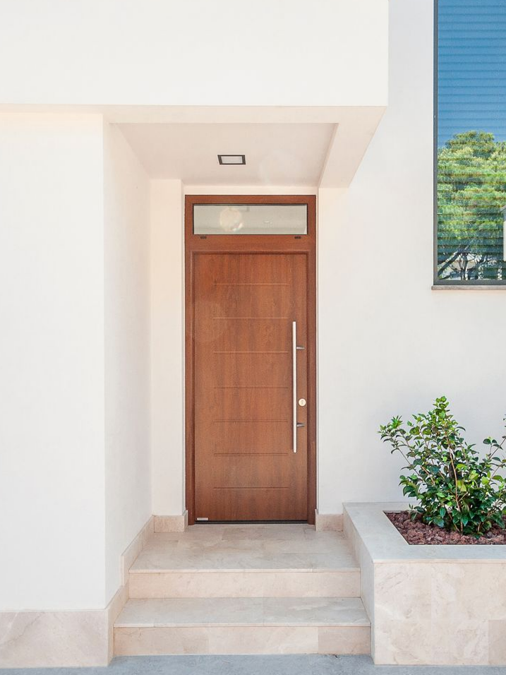 Porte d'entrée en bois avec des lignes horizontales et une petite fenêtre au-dessus, menant à une maison aux murs blancs.