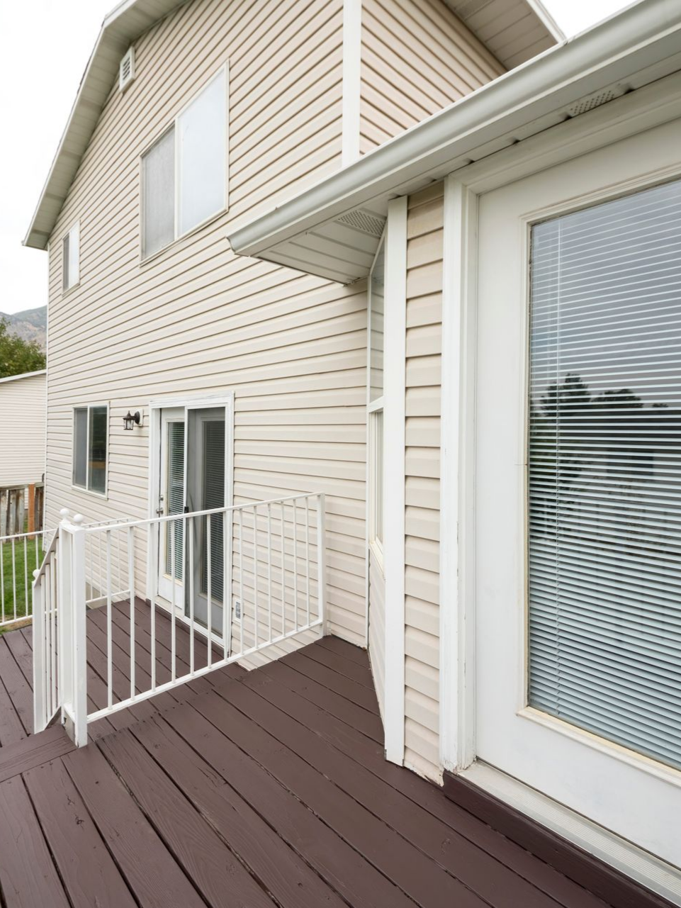Extérieur d'une maison à deux étages avec une terrasse en bois, un revêtement blanc et une porte avec stores.