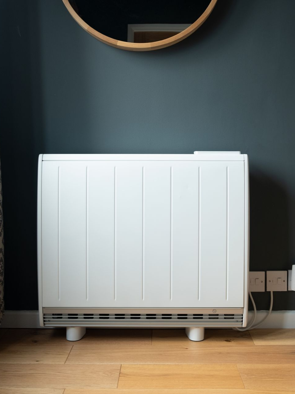 Radiateur électrique blanc sur parquet contre un mur bleu foncé, sous un miroir rond.