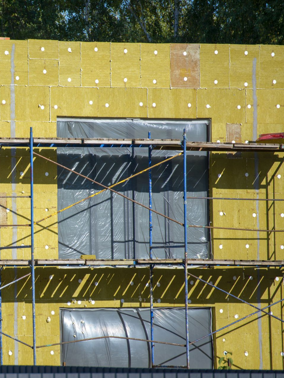 Bâtiment en construction, panneaux isolants jaunes, échafaudage, fenêtres couvertes.