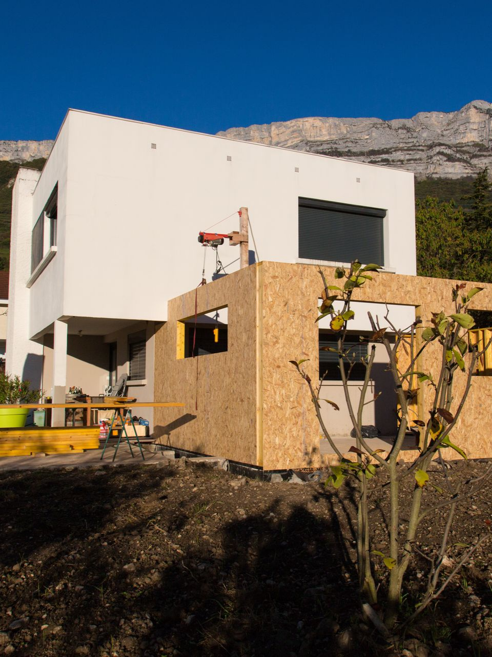 Maison en construction, avec murs en bois devant un bâtiment blanc. Falaise en arrière-plan.
