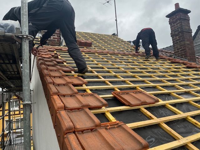 Deux couvreurs installent des tuiles en terre cuite rouge sur un toit avec des poutres en bois et des échafaudages.