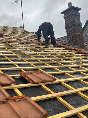 Couvreur installant des tuiles sur un toit résidentiel, avec une cheminée et une antenne visibles sur un ciel nuageux.