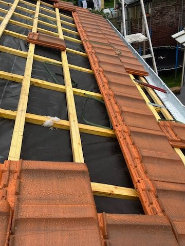 Toiture en construction ; tuiles brunes posées sur une ossature en bois recouverte d'une bâche imperméable noire.