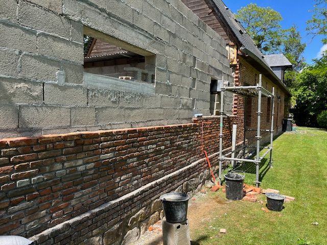 Mur de briques et de parpaings en construction, avec échafaudages et cour en arrière-plan.
