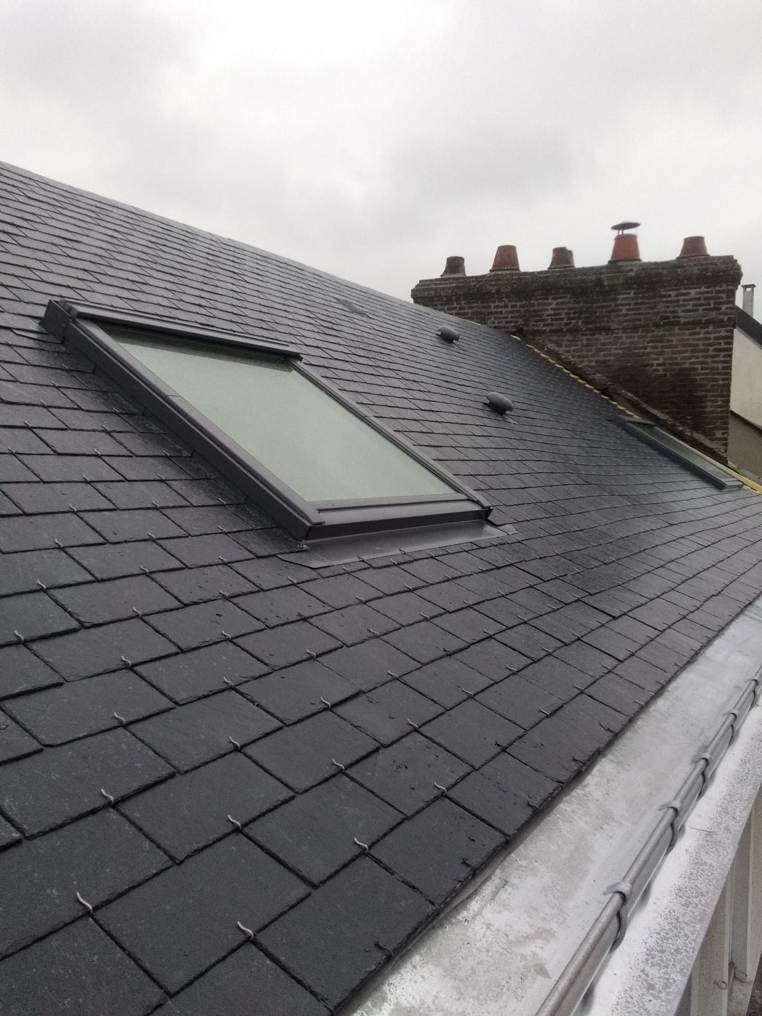 Toit en bardeaux noirs avec puits de lumière et cheminée en briques, journée nuageuse.