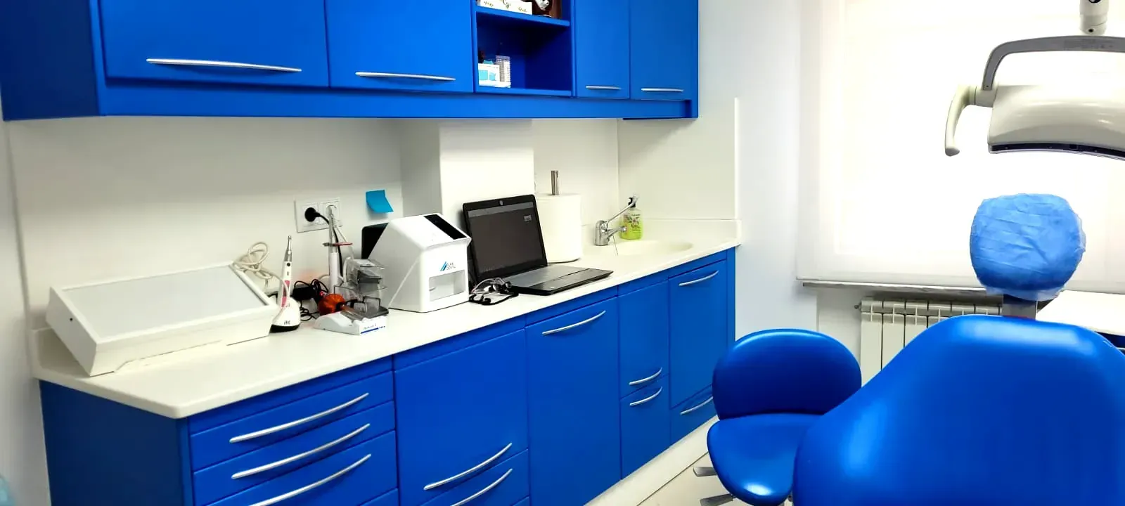 Interior de un consultorio dental. Armarios azules y sillón dental. Encimeras blancas con equipo y una computadora portátil.
