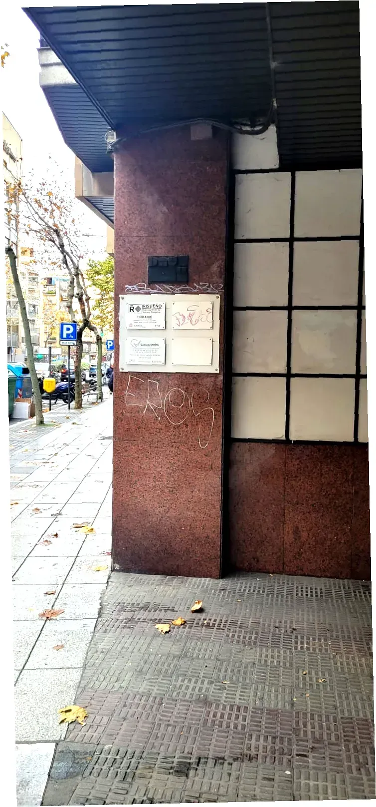 Exterior de un edificio con un pilar rojo texturizado, carteles y una acera a lo largo de una calle.