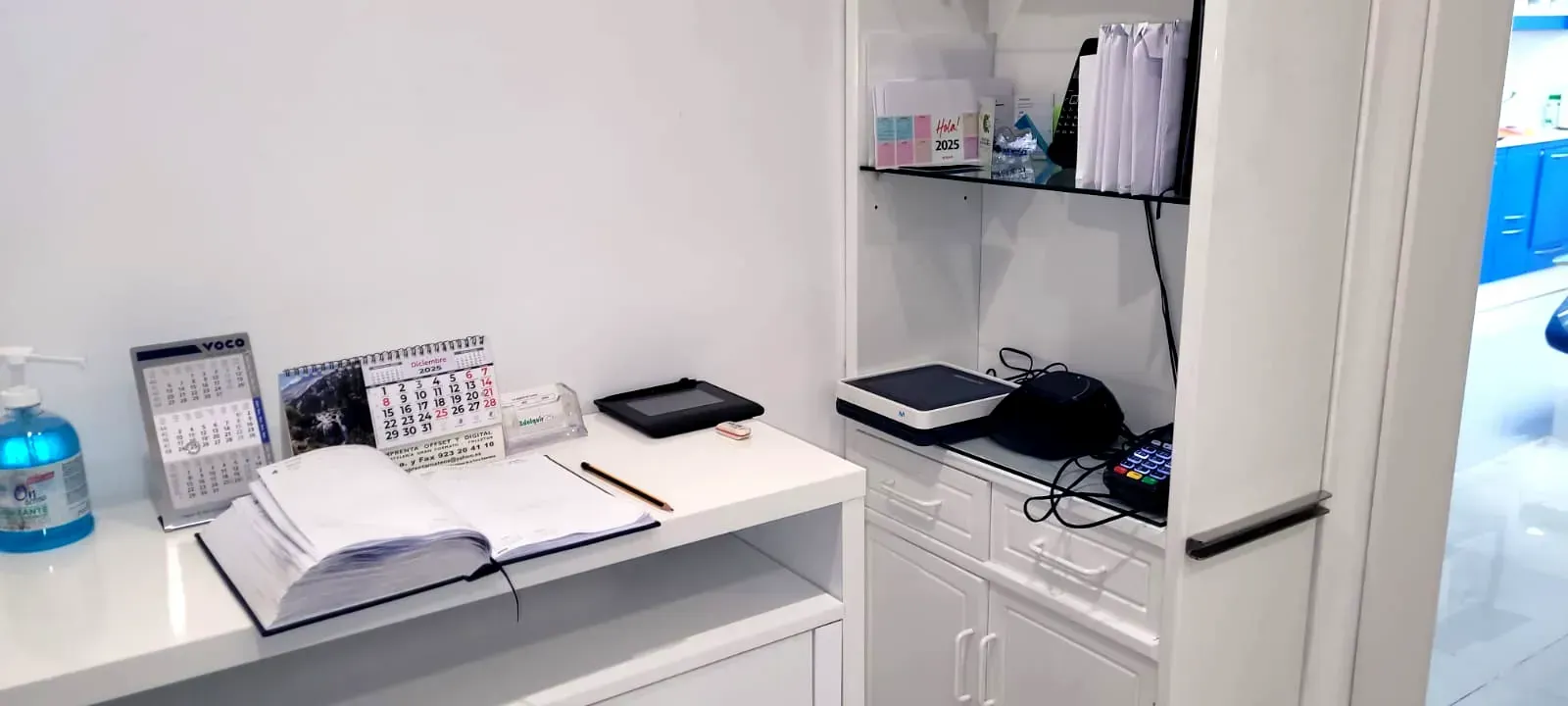 Un escritorio blanco con un cuaderno abierto y calendarios. Se ven estantes con archivadores y una báscula.