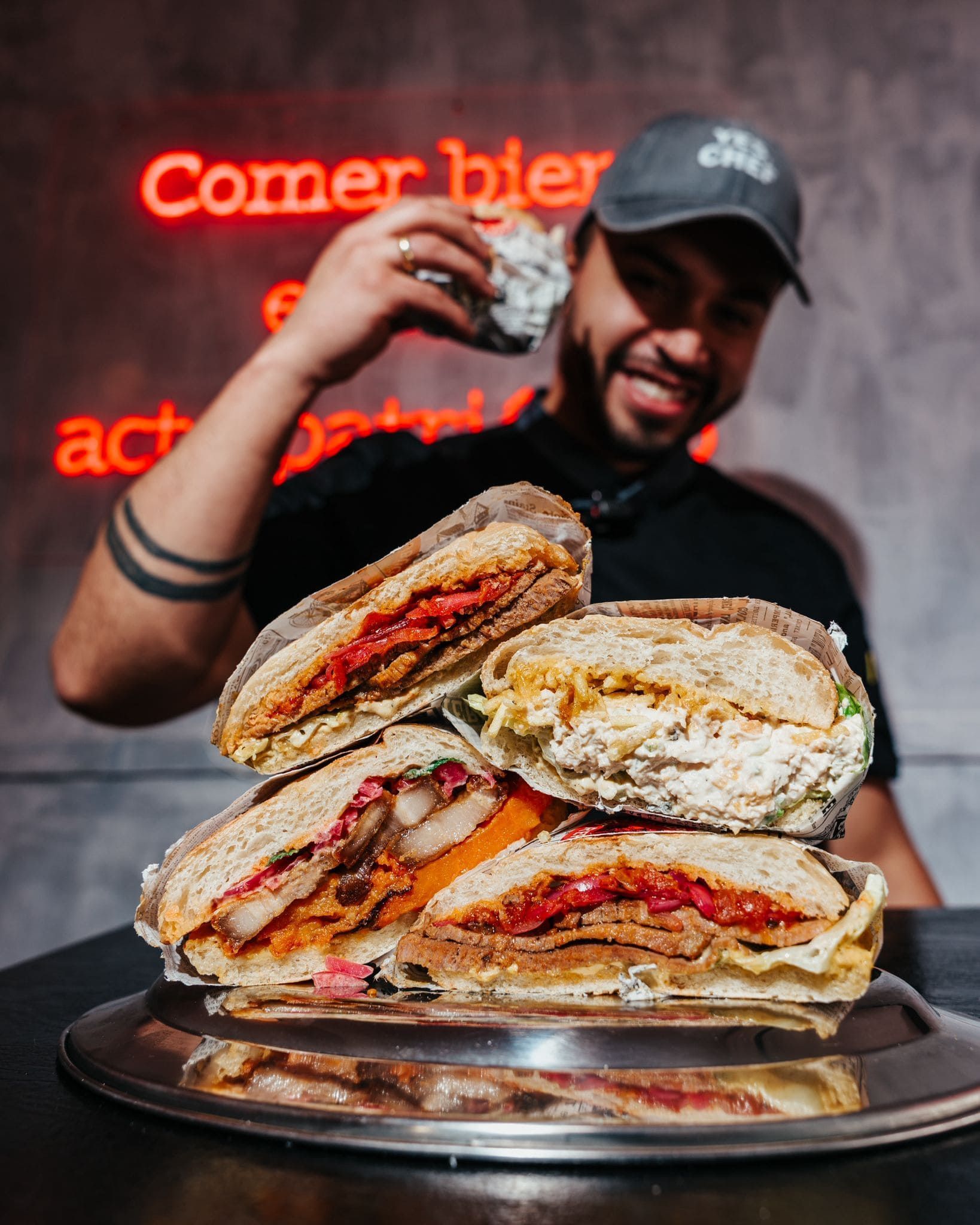 Hombre sonriente con un sándwich en la mano, con una enorme pila de sándwiches en una bandeja de plata delante. Letrero de neón.