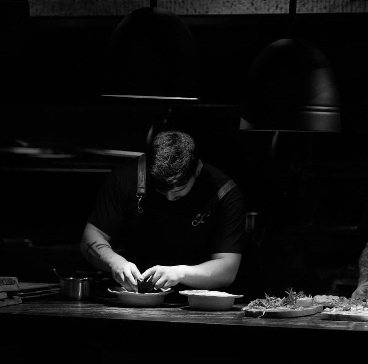 Chef en una cocina poco iluminada, preparando comida en un mostrador, concentrado e iluminado.